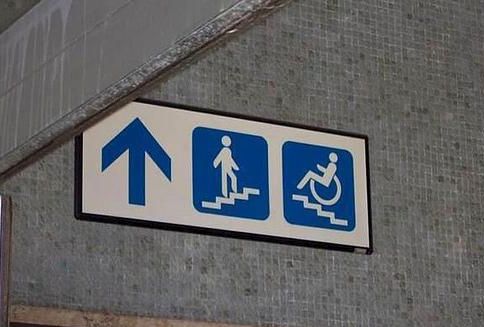 a sign with a arrow pointing up - with a drawing of a person walking up a flight of stairs, and next to that a person in a wheelchair going up a flight of stairs.