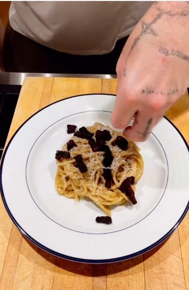 Spaghetti with large burnt squares that look like lumps of shit on a white plate with a hand putting parmesan on top
