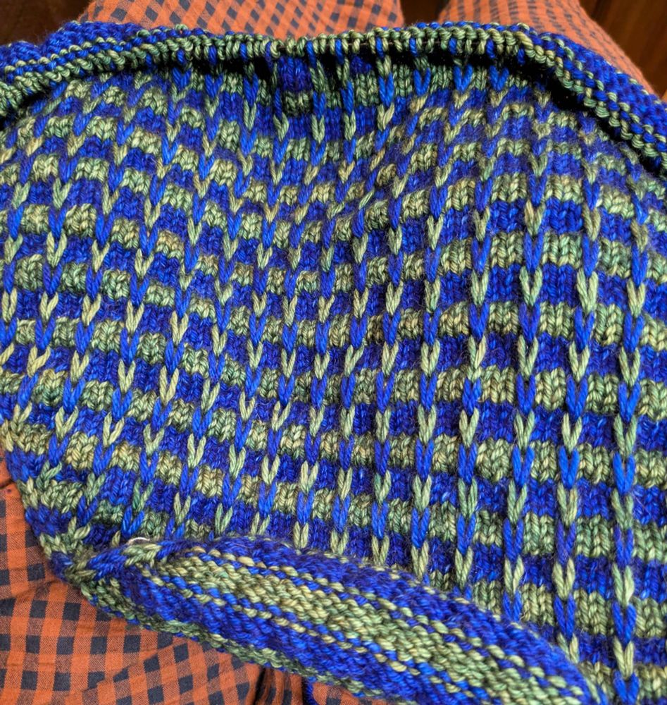 A picture of a piece of knitting in progress, spread out on the lap of a woman in a brown/black gingham dress. The knitting is going to be a cowl to wear around the neck in a cold office, and is blue and green. The blue and green yarns form an almost checkered effect using a combination of plain and slipped stitches. 
