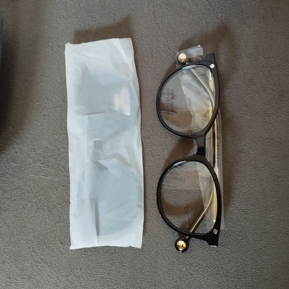 Mostra os óculos fora do estojo e do plástico. Eles são de armação preta, com lentes aparentemente transparentes. Ao lado dos óculos, há o plástico que os envolvia, agora com a lente solar.