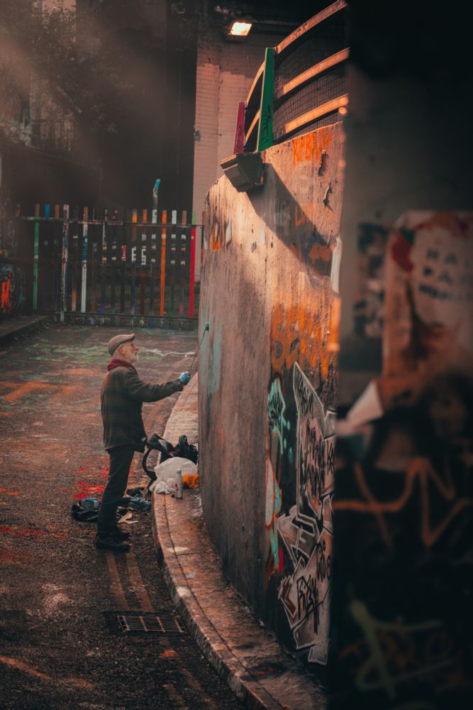 An older gentleman in a flat cap and red scarf paints on a graffiti wall that's dappled in the gentle rays of the winter sun.