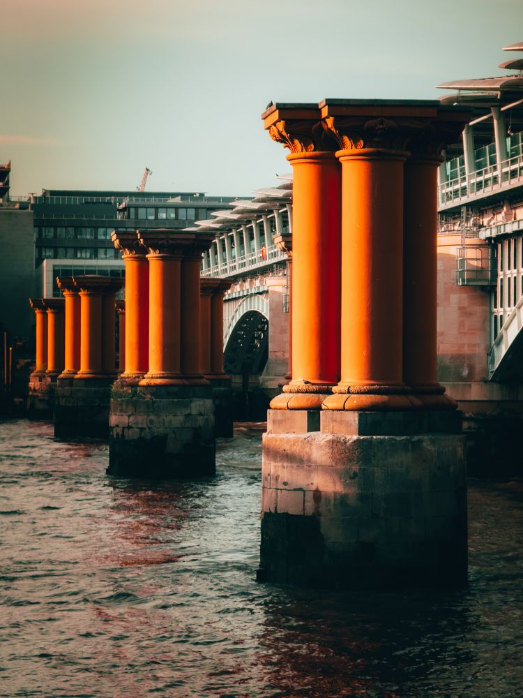 The abandoned pillars of the old blackfriars railway bridge sit at low tide in the Thames lit up golden orange by the late evening sun.