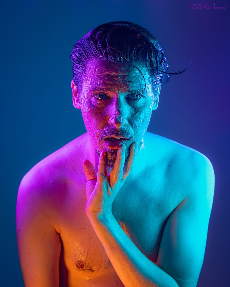 A self portrait by photographer Rab Townsend. The subject is a man with dark, slicked back hair and a goatee, posing topless from the waist up against an indigo background. He is cast in vibrant orange, magenta, and cyan light. A splatter of clear slime is clinging to his face. He is gazing to the right of frame, resting his chin in his left hand, with an expression of puzzlement and sadness.