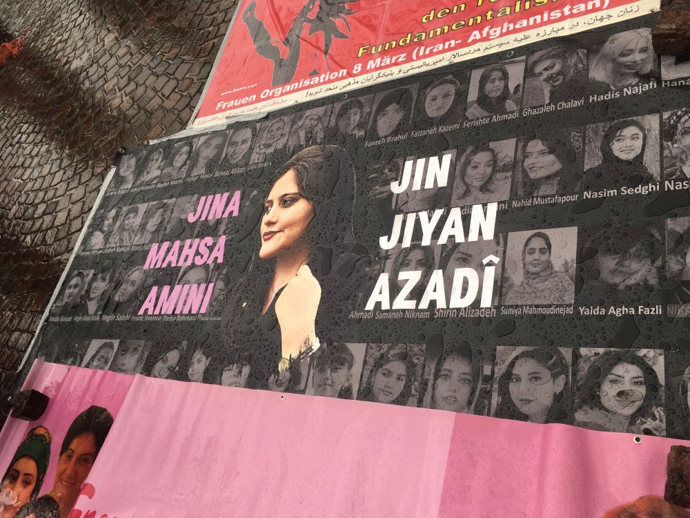 Banner auf der Erde mit dem Bild von Jina Mahsa Amini. Links davon steht ihr Name und rechts Jin Jiyan Azadi