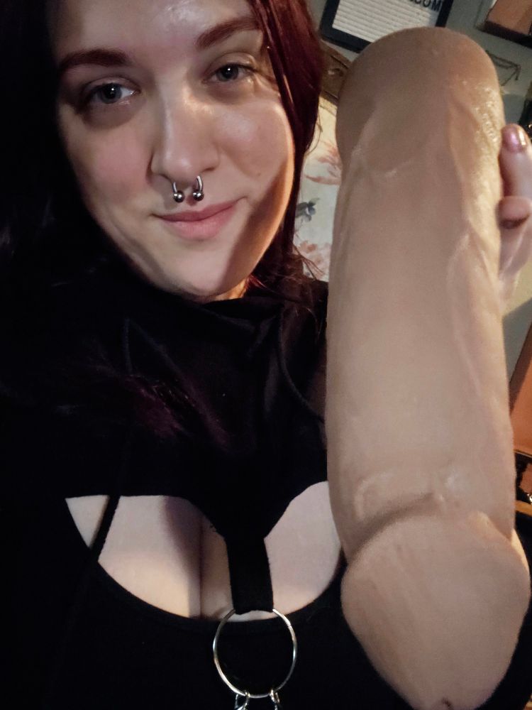a redheaded woman holds up a very large phallus, pointing it at the camera as she smirks.