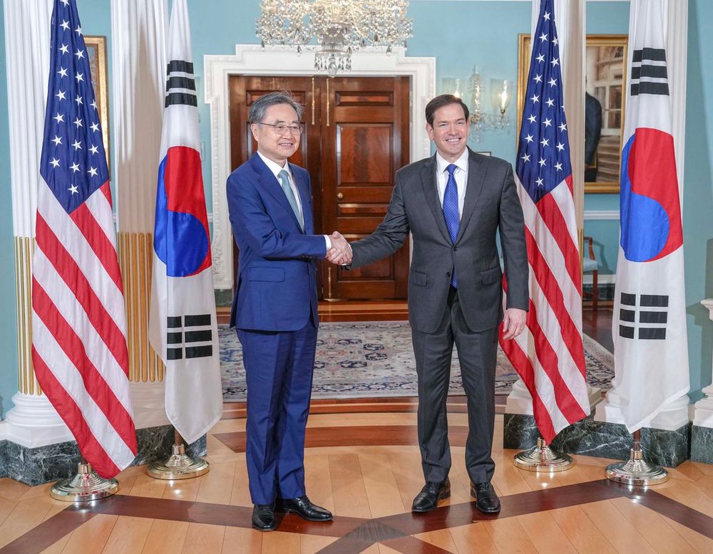 Secretary Rubio and ROK Foreign Minister Cho pose for a photo while shaking hands in Washington, DC on July 31, 2025. U.S. and ROK flags are displayed behind the officials. A chandelier, rug, chair, and painting are visible in the room behind them with a double set of wooden doors. (State Department/Freddie Everett)