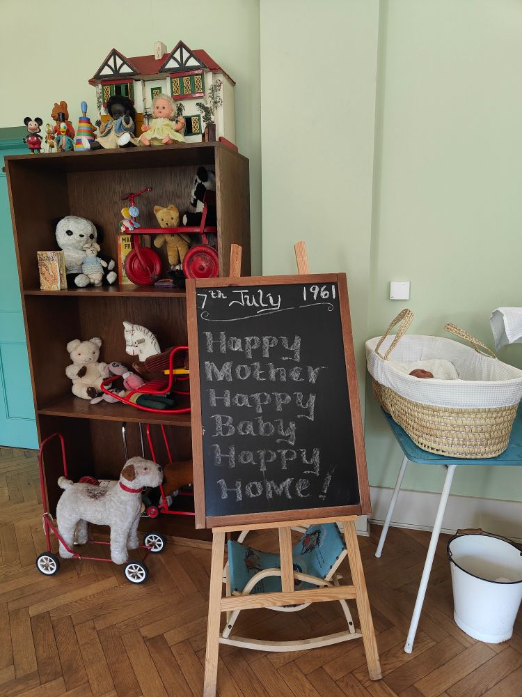 Sign which reads Happy Mother, Happy Baby, Happy Home, and a bookcase filled with children's toys including dolls, a dolls house and push along dogs