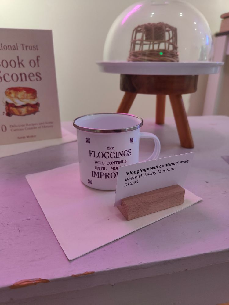 A white enamel mug with 'the floggings will continue until morale improves' in purple. Label with 'floggings will continue mug, Beamish Living Museum, £12.99'