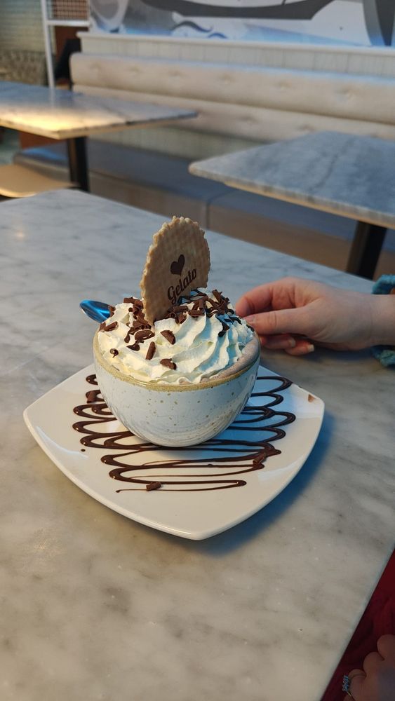 A hot chocolate mug on a grey table. The saucer is drizzled with chocolate sauce, the mug has cream, chocolate sauce, chocolate pieces and a wafer.