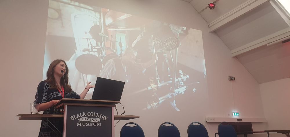 White woman with brown hair and navy dress stands at a lectern, gesturing at an image of a man working a machine with the words "men" and "coal" 