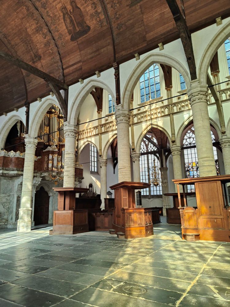 Interior of the Oude Kirk. Plain walls, with a painting just visible on the wooden roof, and tombstone flooring. 