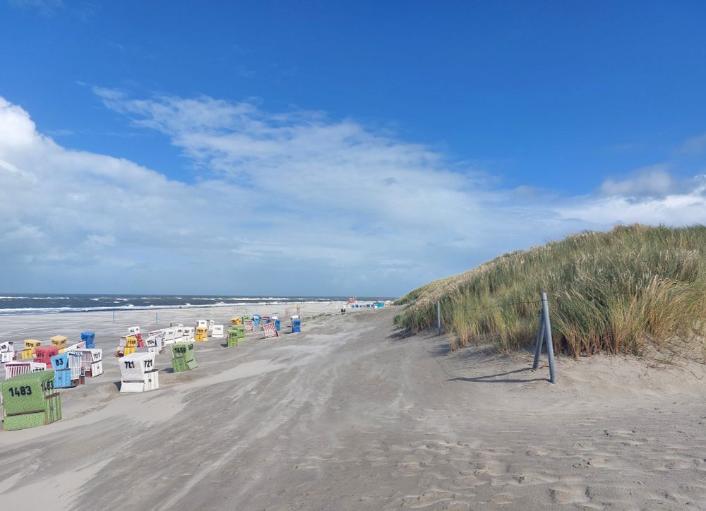 Strand mit Helmgras und Strandkörben. Blauer Himmel mit weißen Wolkenfeldern, Sandverwehungen. 