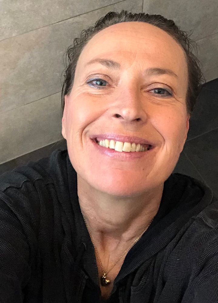 Frau Mitte 50 steht schwarz gekleidet im grau gefliesten Bad mit nassen Haaren und lächelt glücklich und hoffnungsvoll ins von oben aufgenommene Selfie.