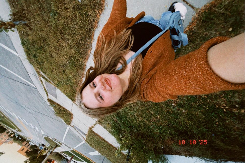 a selfie of jori walking on a sidewalk, she is wearing an orange cardigan, black tank top, and jeans