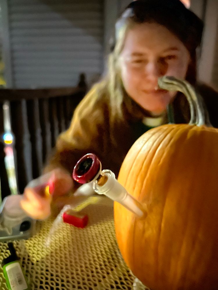 a pumpkin made into a bong on a table, jori is suffering behind it bc a lot of smoke can fit in that thing