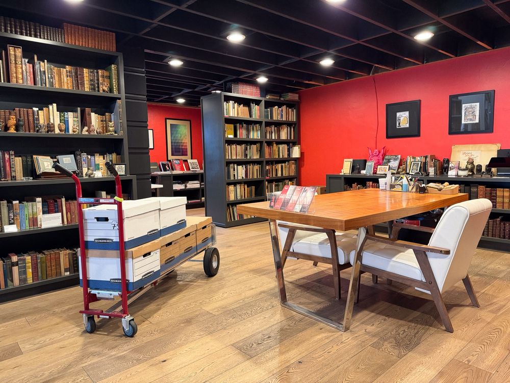 A cozy bookstore area with bookshelves, a red hand truck with storage boxes, a wooden table, and white chairs.