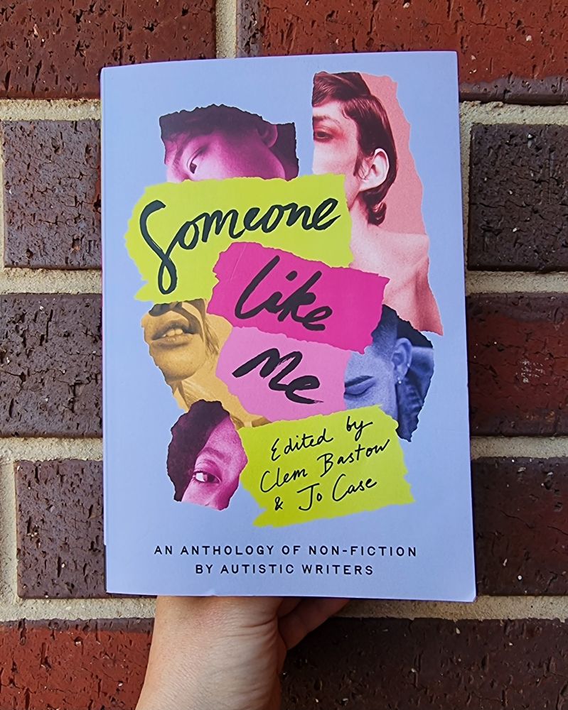 A handheld book of 'Someone Like Me' against a background  red brick wall 
