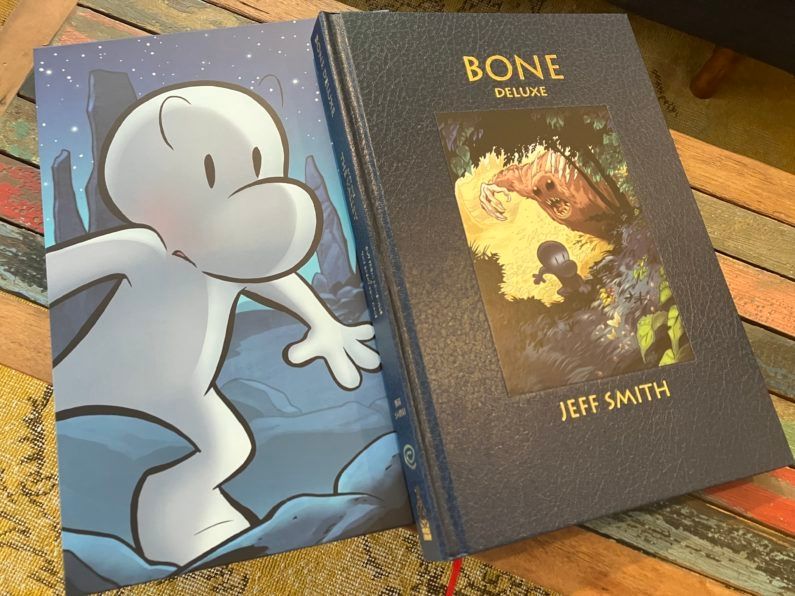 Bone: The Deluxe Edition vol. 1 by Jeff Smith
A hard cover book lying on top of its slipcase.
The book has a dark blue leather effect Cover with a picture inset into it. The picture is of a small, white, human-ish, very cartoony figure with a big nose (Bone) running through a very detailed forest and being chased by large, brown, furry creature with black eyes and its mouth open wide showing lots of big, sharp teeth. One clawed hand is raised as if about to strike.
The slipcase is a full figure drawing of Bone at night. He looks like he is creeping through some mountainous terrain and looks worried. 