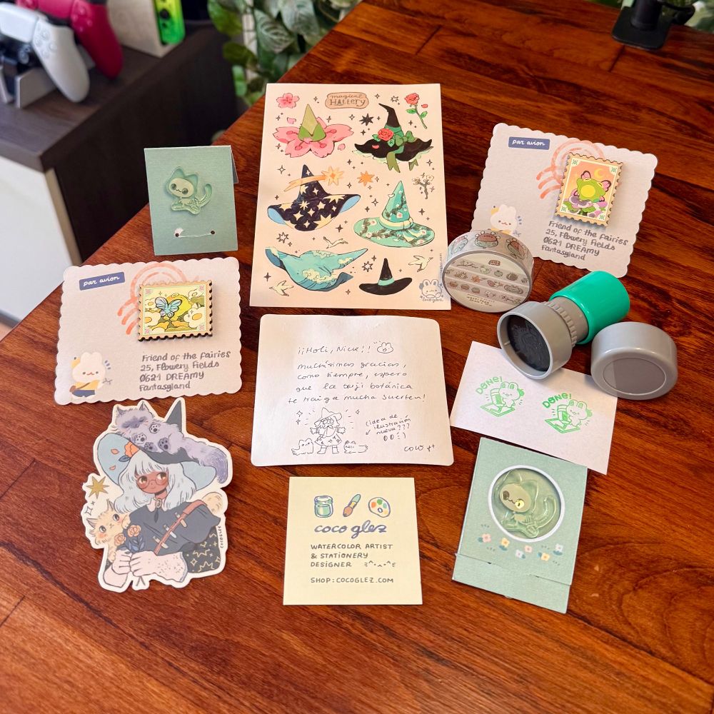 Foto de ítems de la tienda de la artista cocoglez:
-Una lámina de pegatinas de diversos sombreros de bruja florales. 
-Un par de pins acrílicos de gatetes esqueletos muy adorables. 
-Un par de pins de madera de sellos postales de una mariposa y una rana. 
-Un cacharrito para estampar sellitos de tinta con una ranita diciendo “Done!”.
-Un rollo de washitape de elementos otoñales con bellotas y hojitas. 
-Una tarjetita de visita. 
-Una notita de agradecimiento muy linda escrita a mano. 
-Y para acabar siempre una pegatina de la brujita más Nickcore ever: pelito clarito en contraste con su color oscuro de piel, gatitos, flores y estrellas. 🥰