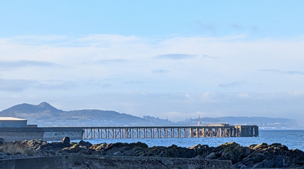 A distant blue hill to the left, with buildings just visible on its lower slopes to the right. An industrial jetty running out into an icy blue sea in the mid-ground. dark rock shore in front. Light flat cloud and a cold pale blue sky above.