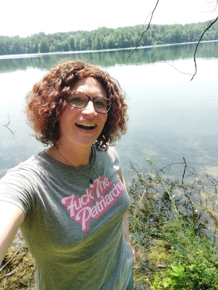 A smiling woman with glasses and short-ish curly hair standing in front of a large body of water. She wears a shirt that says "Fuck the Patriarchy".