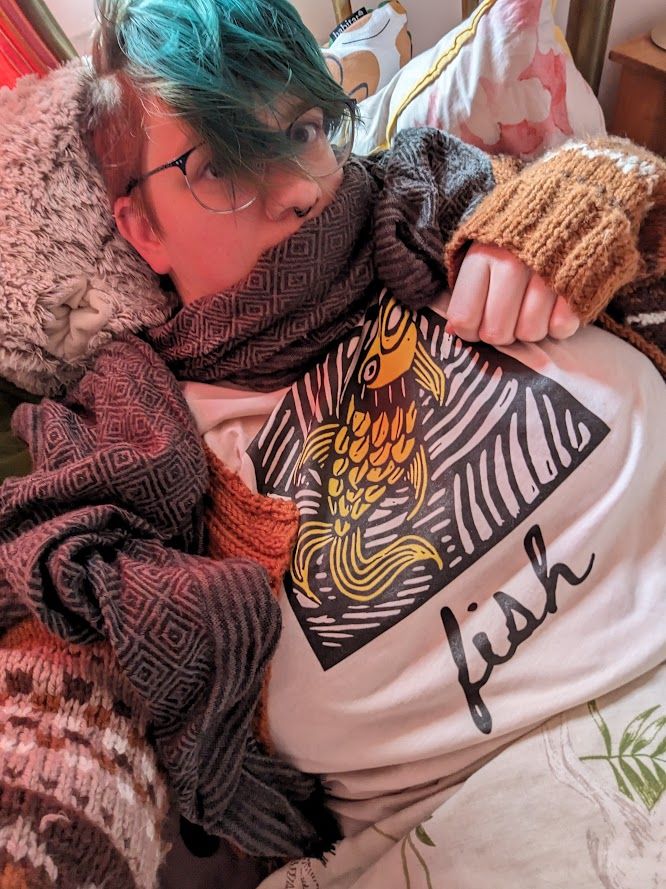A selfie of me wearing a white shirt with a yellow and black illustration of a particularly gormless-looking fish, captioned in cursive with the word "fish". The art is from the Fish card of Latimer's Lenormand, a Lenormand divination deck by Nell Latimer of Unquiet Arcadian