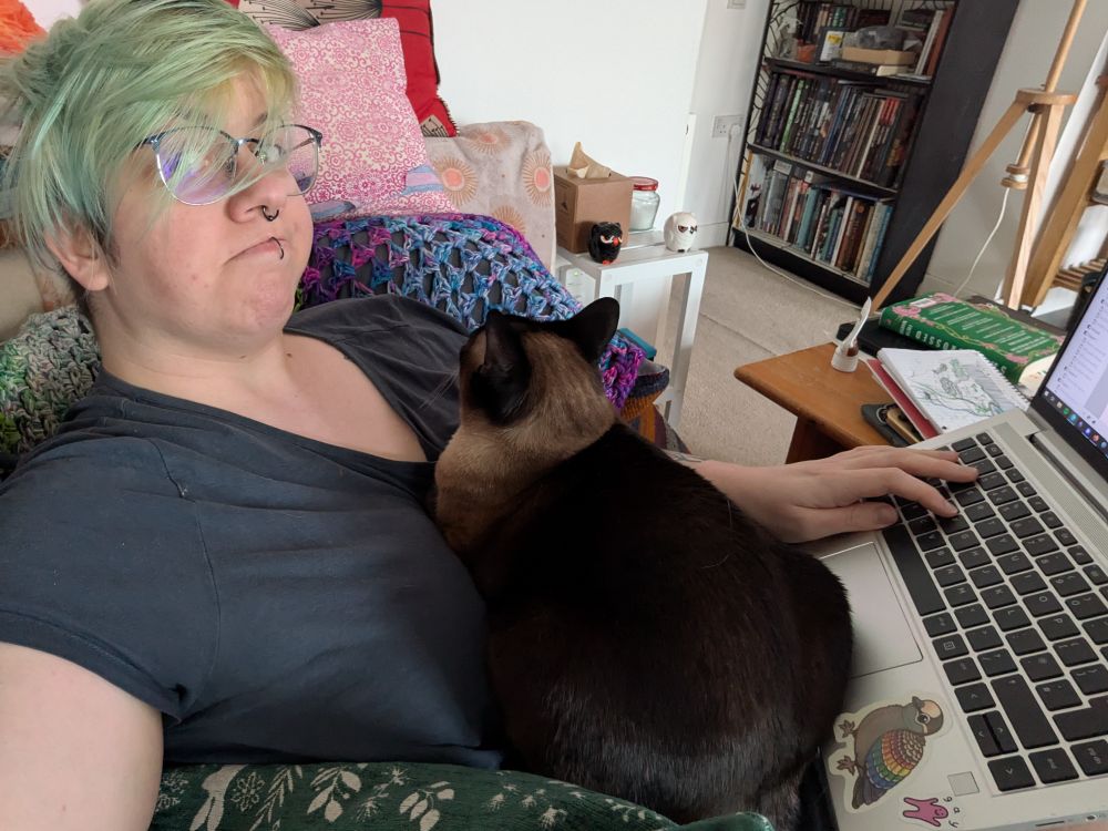An example of my seal point Siamese cat, Ishtar, in her "nurse Ishtar" mode. The photo is a selfie of me (a white person with green hair and glasses) laying on my sofa with a computer on my lap. This was a day when I had a bad long covid flare-up. Ishtar has firmly wedged herself between the computer and my face, and she's loafed up very compactly on my chest. Her ears are up and alert and she's looking directly at me, monitoring the unruly patient that keeps insisting on doing things instead of resting.