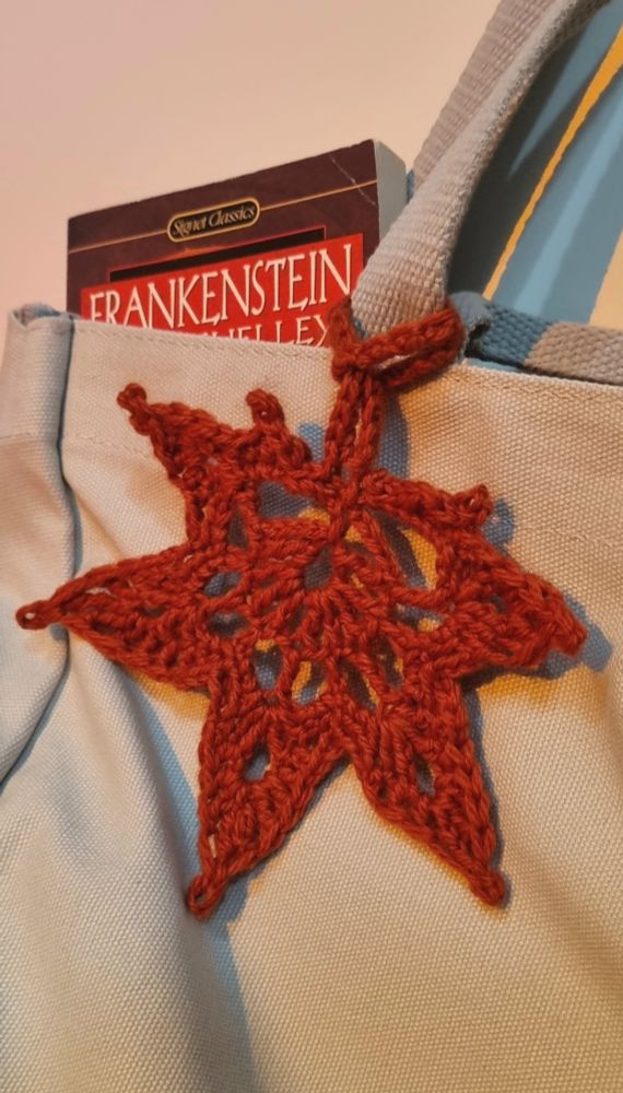 a dark orange crocheted maple leaf bag charm wrapped around the straps of a grey tote bag. a copy of Frankenstein by Mary Shelley peeks from the tote bag. the leaf references the movie Frankenstein (2025) directed by Guillermo del Toro.