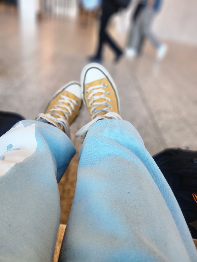 Picture of legs in blue sweatpants and yellow converse shoes stretched out from the ledge of a bench while people walk by in an airport. 