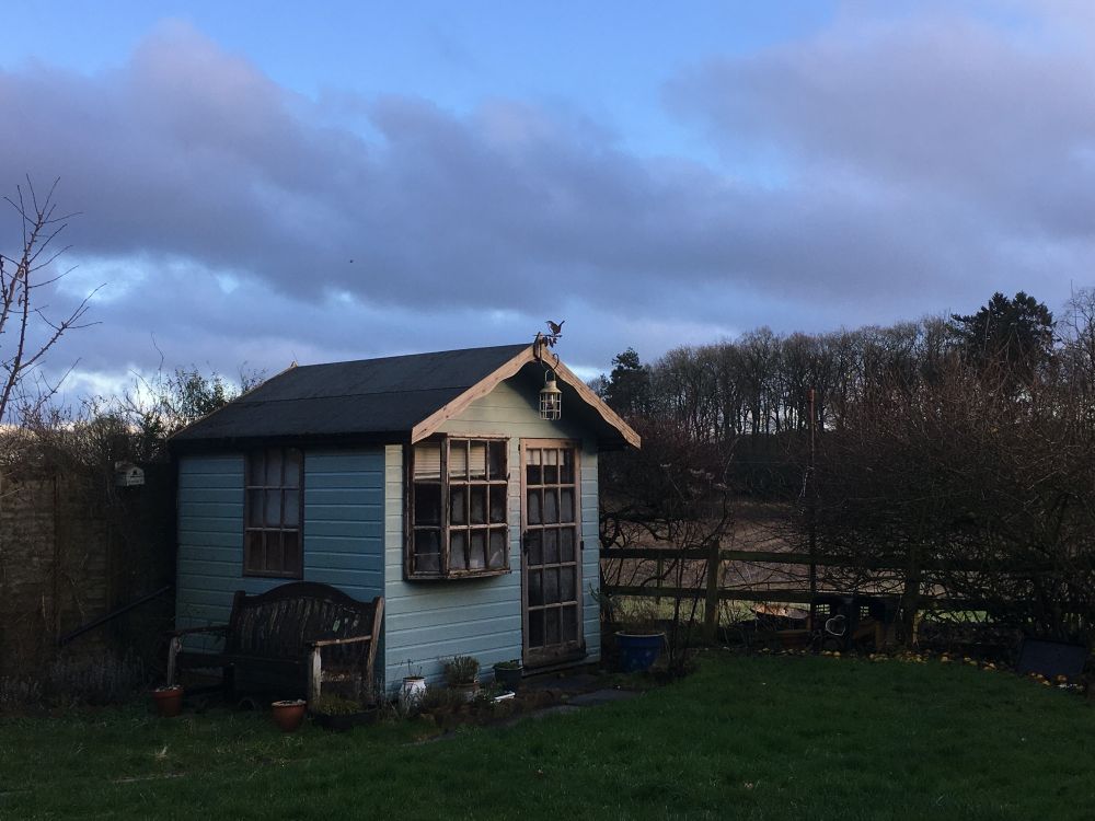 Little blue bow-windowed hut in a garden backing into fields and a wood