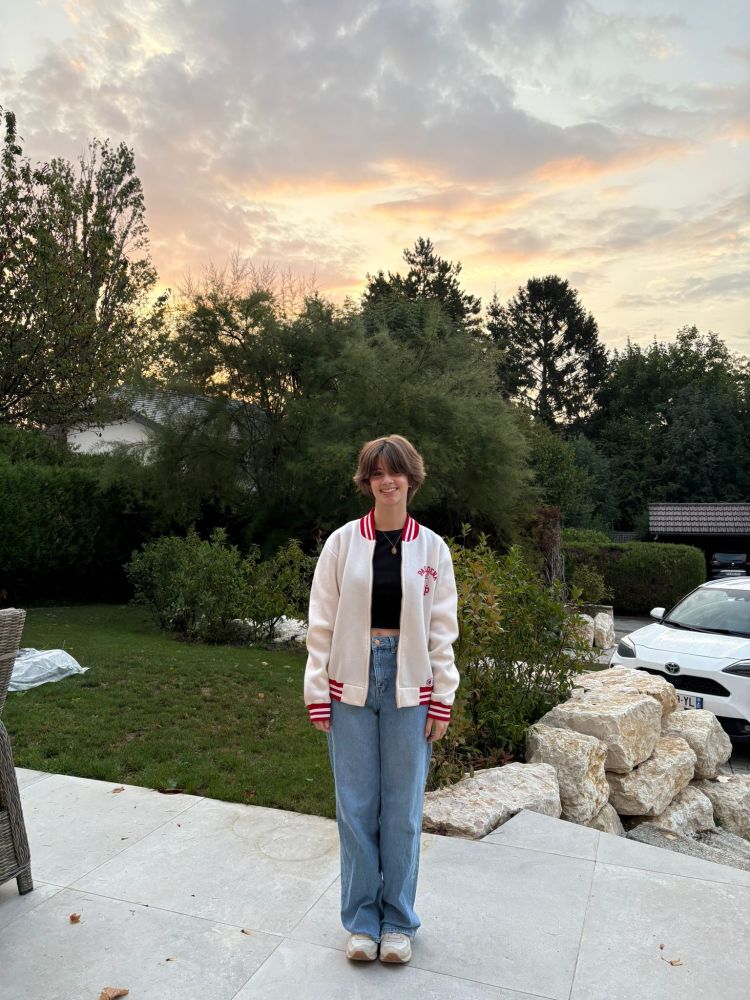 Our 17-year-old daughter stands on our front patio with our front yard and a pink and gold-tinged sunrise in the background. She's wearing jeans, a black top, and a white and red varsity jacket. She has short brown hair and a big smile. 
