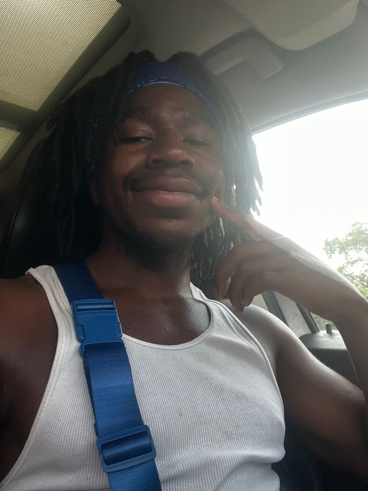 Selfie of a man in a car with a white tank top and blue strap over his shoulder 