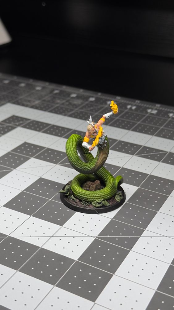 Painted miniature of a coiled green snake with an open mouth, featuring a warrior riding on its back and wielding a flaming weapon, placed on a grid-patterned surface.