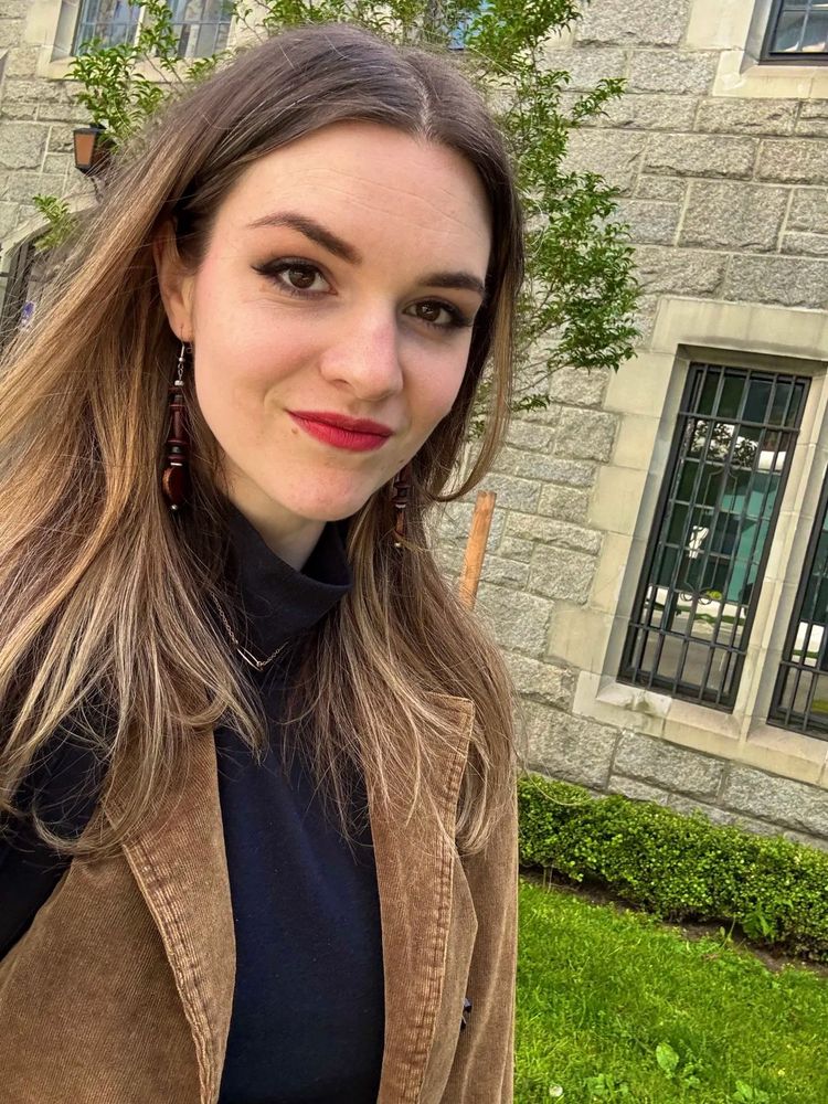 A selfie of me, Sarah Z! A white woman with long brown hair that fades to blonde at the ends, wearing a black turtleneck, brown corduroy jacket, bold red lipstick, and dark eye makeup. She has dangling earrings and is smiling slightly. The background shows a stone building with barred windows, green grass, a small hedge, and a tree with green leaves.