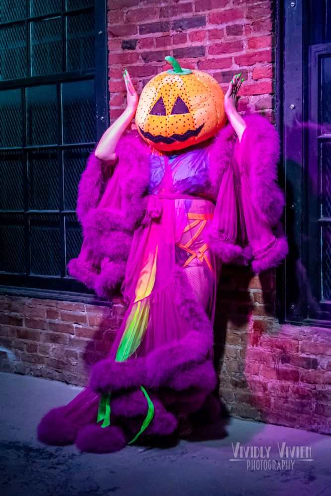 A very vibrant apparition of a burlesque performer stands in front of a brick wall with windows, in a searingly vibrant violet feather-trimmed robe with a hint of silks in neon colors and strappy orange lingerie underneath.
The performer’s head is covered by a giant jack-o’-lantern head.