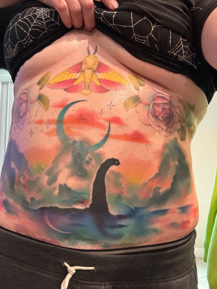 A photo of a torso covered in tattoos. The sternum features a rosy maple moth, flanked by two pastel pink and blue roses and honeysuckle bunches. Beneath that on the belly is a vista of three mountain peaks with a giant crescent moon rising behind the middle peak. Coral clouds and colorful mists swirl around the mountains, while the Loch Ness Monster swims in a pool in the foreground.
Friends have described this scene as a Lisa Frank cryptid watercolor.