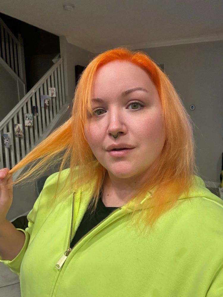 A brightly-colored nonbinary creature stands in a high-vis yellow hoodie with a black tee underneath. They are drawing attention to their freshly colored hair, which is intense orange at the roots and is a slightly less intense peachy orange along the rest of the length at their shoulders.