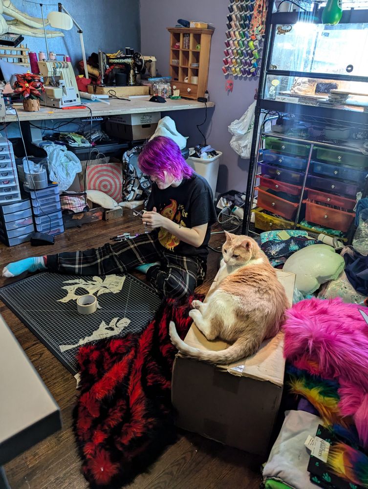 A person sits on the floor of a crowded workroom working with patterning tape while an orange cat looks on from his perch on a box.