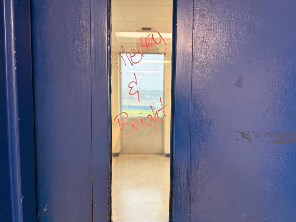 Rectangular door window looking to an enclosed corridor & exterior window. The door's window has "Merry & Bright" written in red on it. 