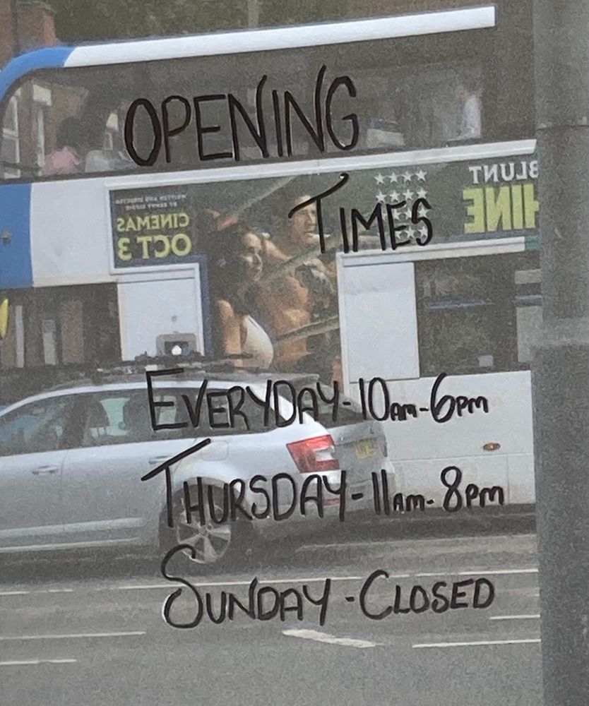 A shop’s opening hours written on a glass door that is reflecting the street. The hours read:
“Everyday 10am-6pm
Thursday 11am-8pm
Sunday - Closed”