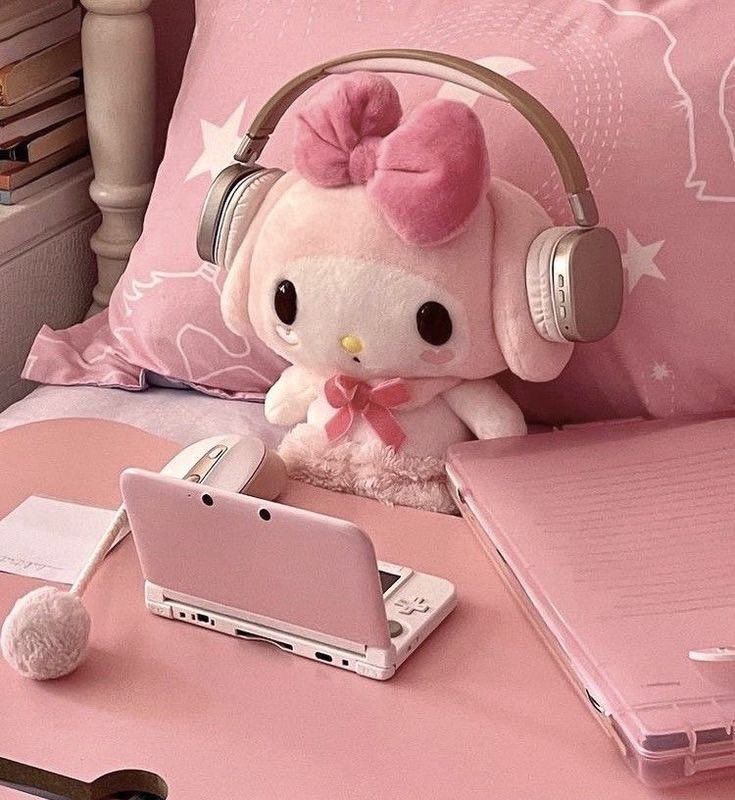 my melody (pink bunny) plushie with headphones on, looking at a pink and white 3ds! shes surround by pink items, like a pink bedding, pencil, and pink case next to her!