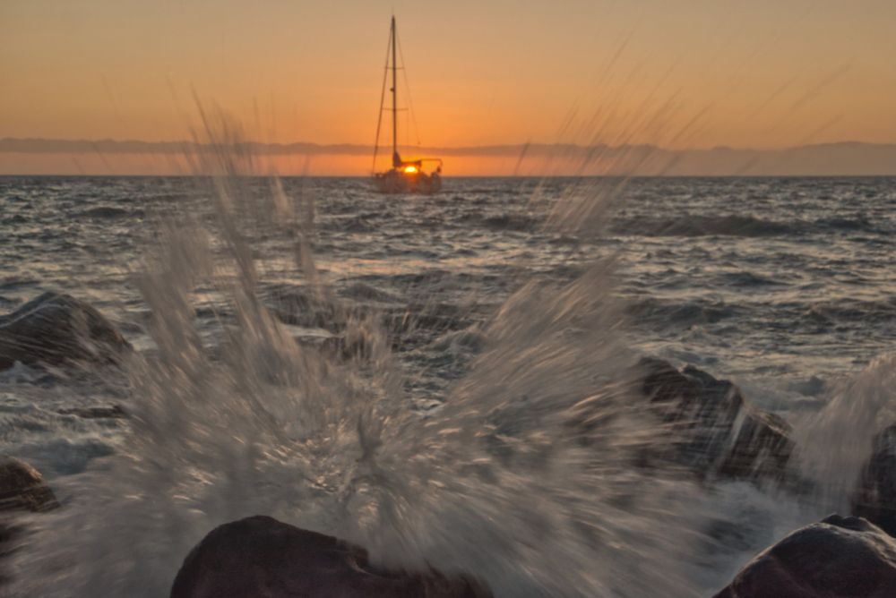 A wave splashing on rocks, beyond a yacht is moored for the evening and the sun is settling directly behind it.