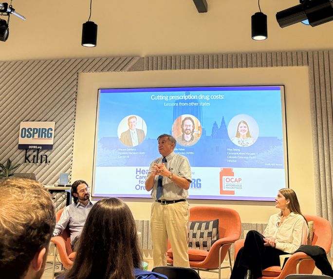 Photo of a room, backs of two folks heads are visible. At the front three people are in front a slide show.