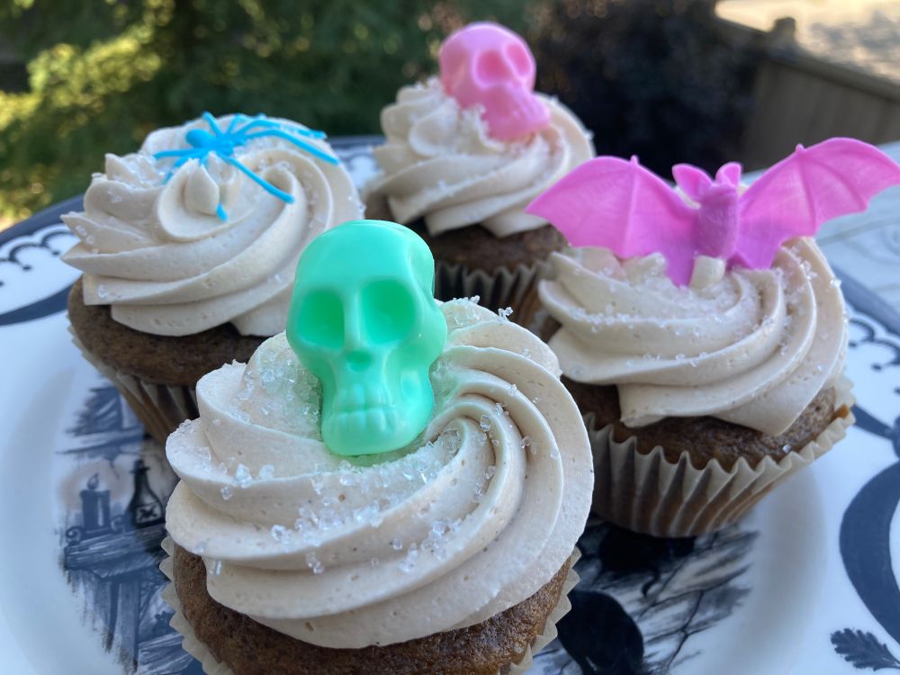 four cupcakes on a plate. the cupcakes are dark brown from steeped earl 
grey tea, with piped cream coloured vanilla bean frosting. they are decorated as follows- one mint green plastic skull, one pink plastic skull, one blue plastic spider, one pink plastic bat.