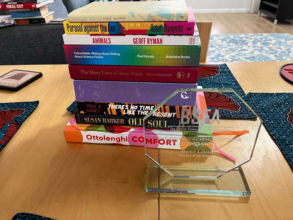 A pile of books purchased on a recent trip. From to to bottom: Houses of Ravicka by Renee Gladman, Parasol Against the Axe by Helen Oyeyemi, Animals by Geoff Ryman, Colourfields by Paul Kincaid, The Many Lives of Anne Frank by Ruth Franklin, The Raven Scholar by Antonia Hodgson, There’s No Time Like the Present by Paul B. Rainey, Old Soul by Susan Barker, and the cookbook Ottolenghi Comfort. In the foreground, a trophy for the 2024 BSFA award for long non-fiction.