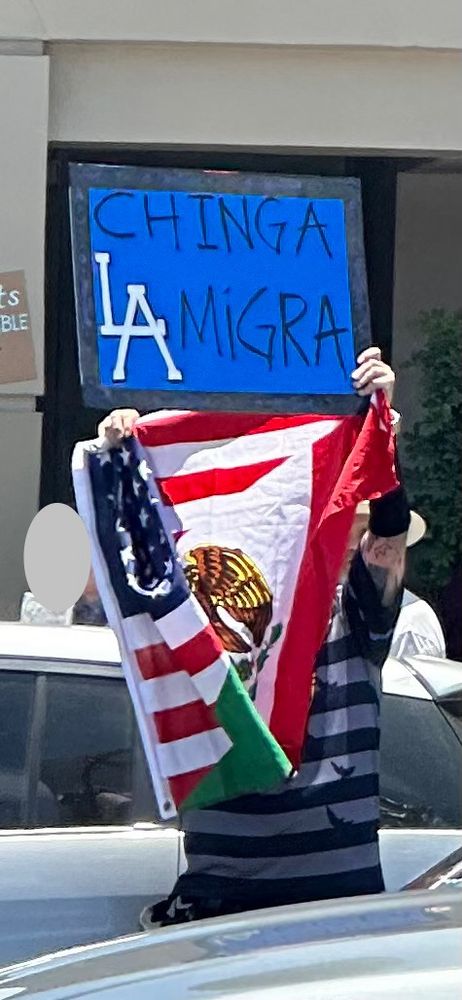 A man holds up a sign saying “Chinga La Migra” and a Mexican flag. The LA resembles the dodgers emblem