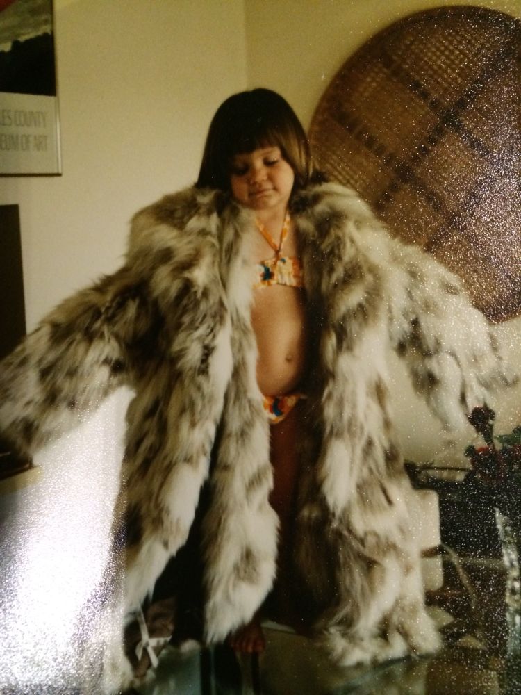 A child in a bikini with an adorable pot belly and a brunette bowl cut stands on a table wearing an adult woman’s fur coat.