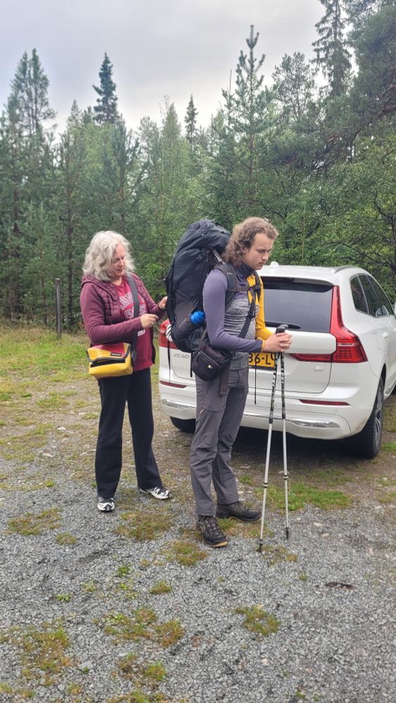 Bosomgeving. Jongeman met grote rugzak en twee nordic walking sticks staat klaar voor vertrek. Zijn moeder verschikt nog iets aan de rugzak. Achter hen een geparkeerde witte Volvo.