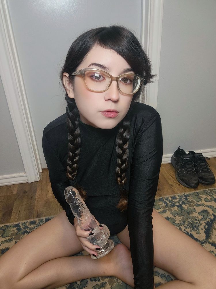 Dark-haired girl with glasses and two long braids sitting cross-legged on the floor holding a clear dildo.
