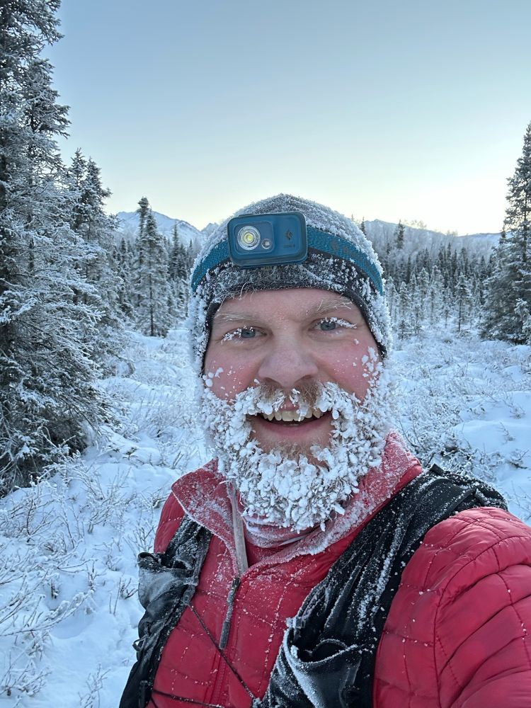 Runner with a frozen icy beard. Snowy trees and mountains in the background. Clear blue sky. 